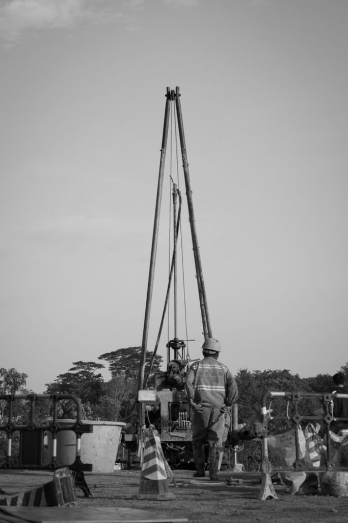Black and white photo of a worker supervising drilling equipment outdoors.