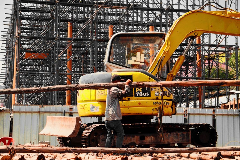 A construction worker manages logs with an excavator at a busy industrial site with scaffolding.